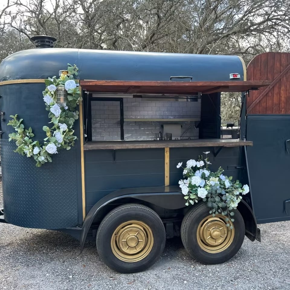 Dark blue food truck with gold trim, open serving window, and white floral decorations.