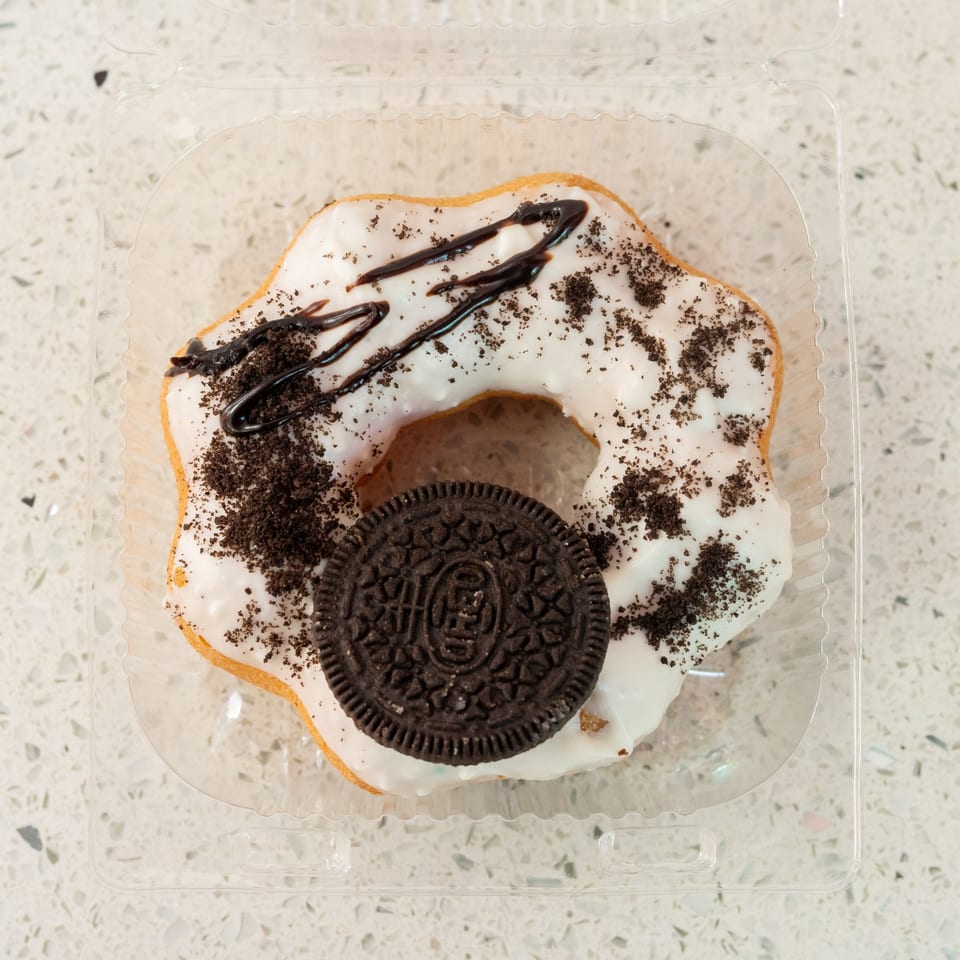 White-frosted donut with chocolate drizzle, cookie crumbles, and an Oreo in a plastic container.