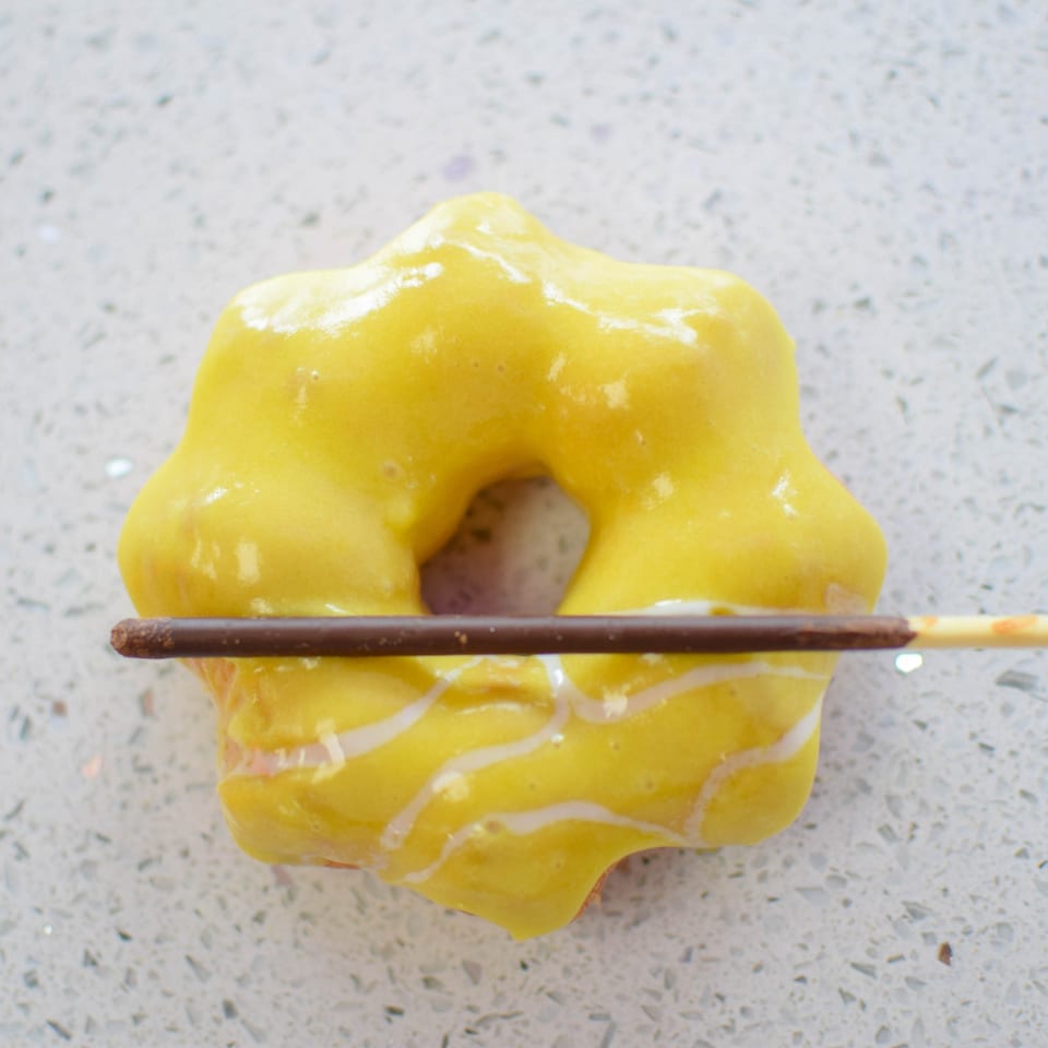 Yellow glazed donut with white icing stripes and a chocolate stick through its center.