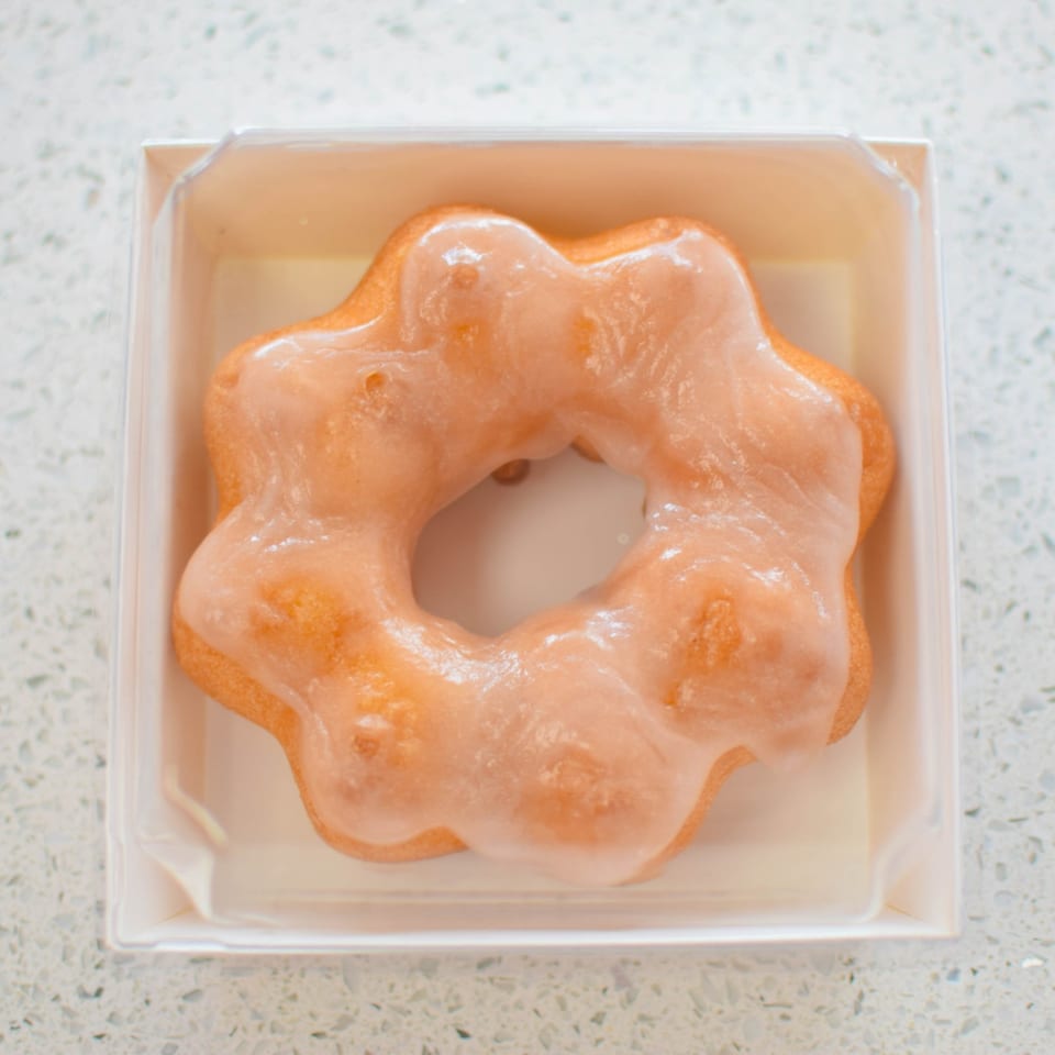 A glazed mochi donut, shaped like a flower, rests in a white paper box.