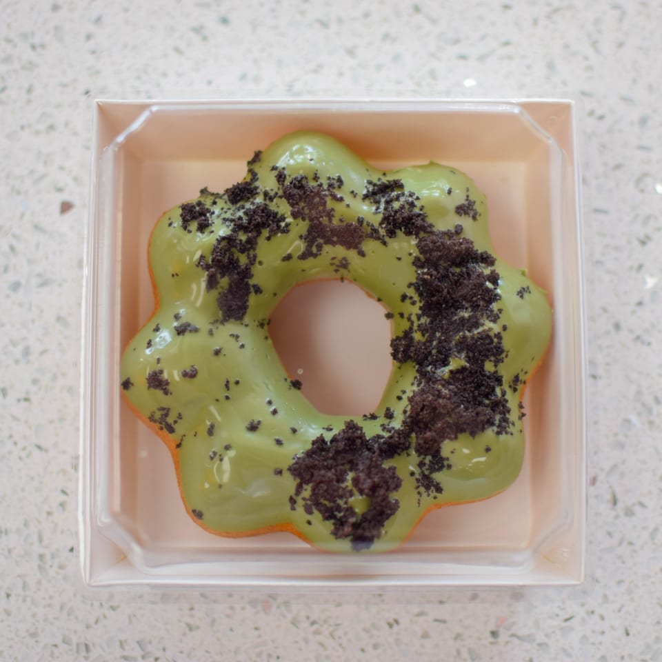 A mochi donut with green matcha glaze and chocolate cookie crumbles in a clear plastic container.