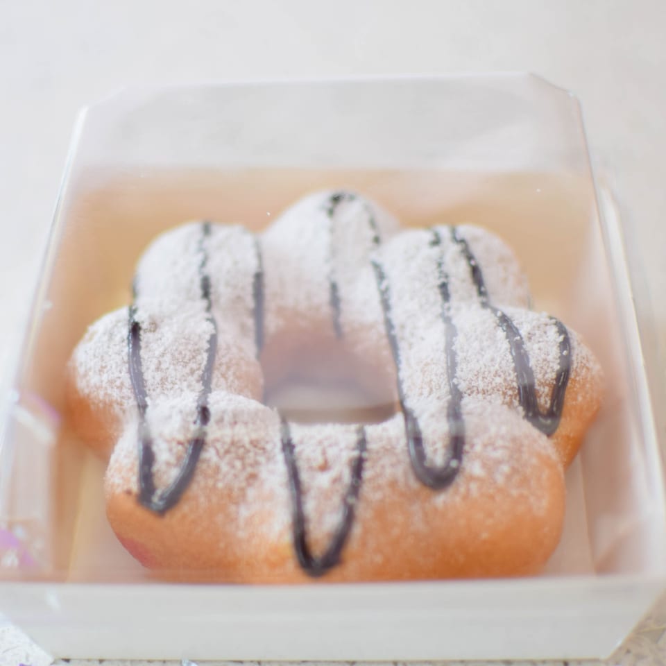 A mochi donut, dusted with powdered sugar and drizzled with chocolate, rests in a clear plastic container.