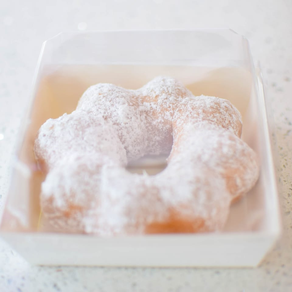 A mochi donut, shaped like a flower, covered in powdered sugar, sits in a white box.