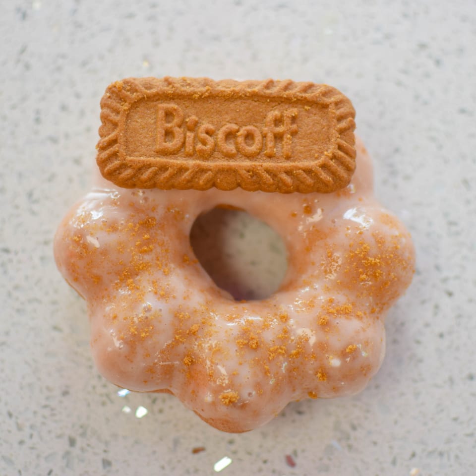 A Biscoff cookie sits atop a glazed, flower-shaped donut sprinkled with cookie crumbs.