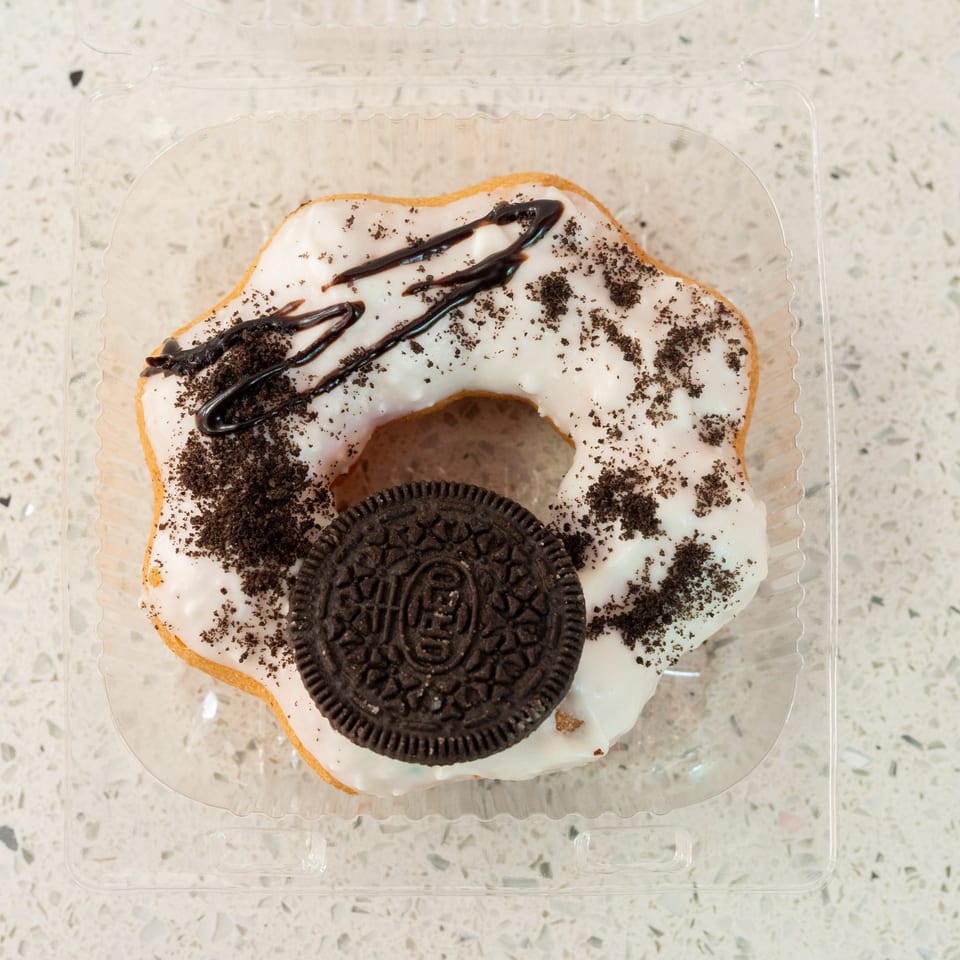 A white-frosted donut with chocolate drizzle and cookie crumbles, topped with an Oreo, in a plastic container.