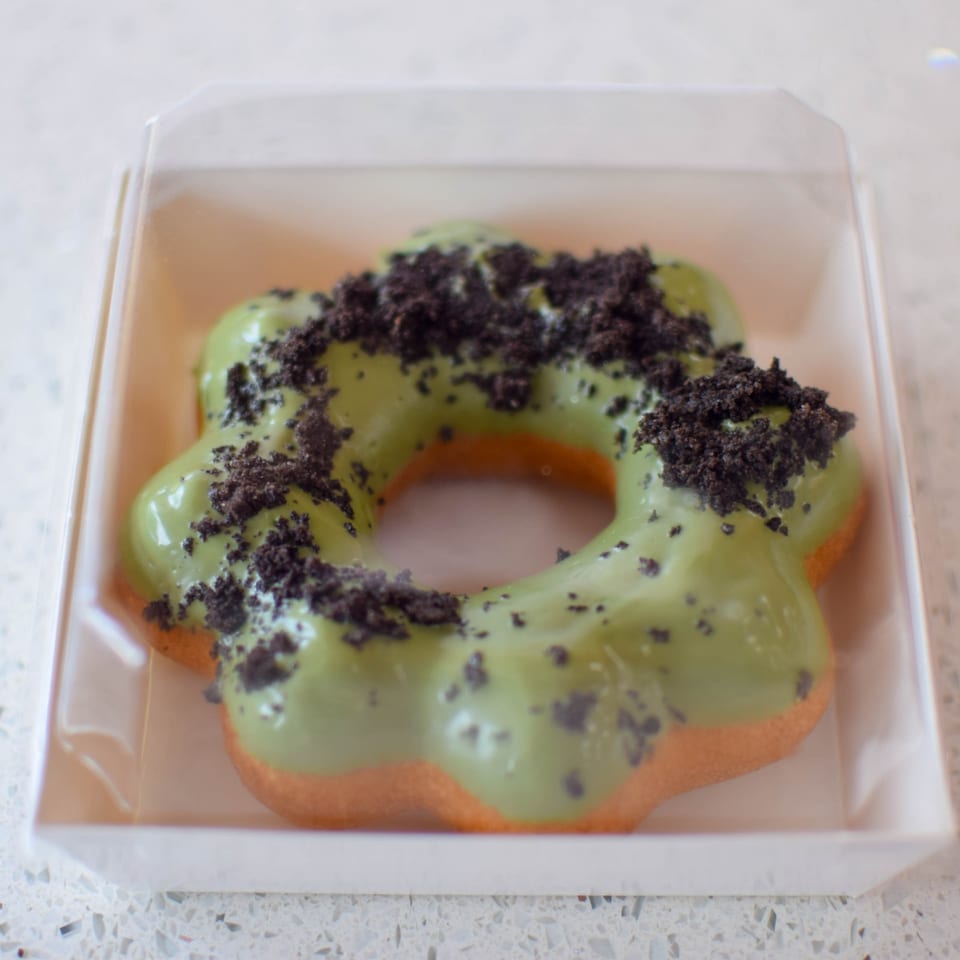 A flower-shaped donut with green matcha glaze and dark cookie crumbles in a clear plastic box.