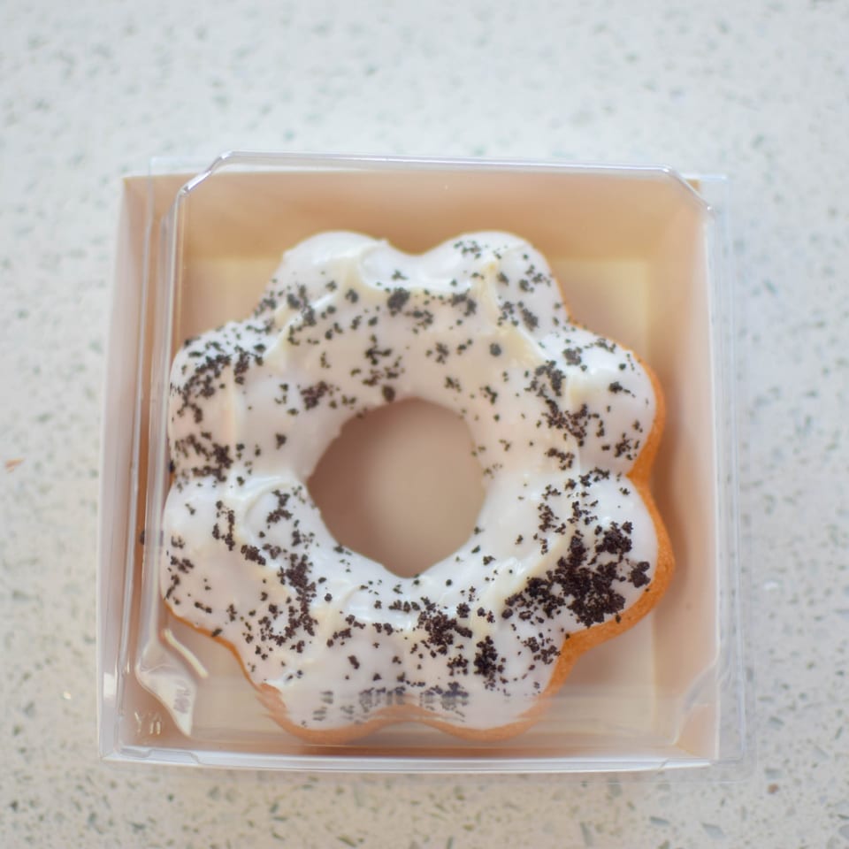 A flower-shaped mochi donut with white glaze and cookie crumbles in a clear plastic container.
