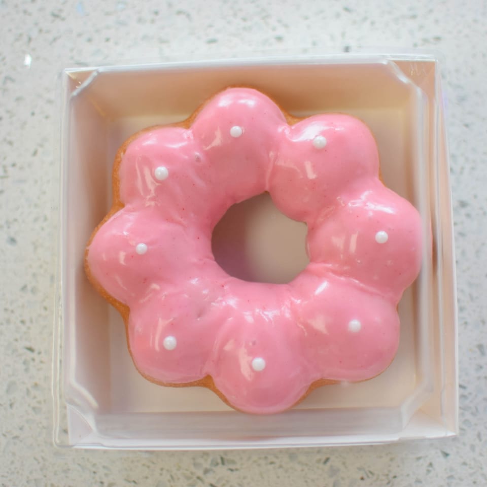 Pink mochi donut with white sprinkles in a clear plastic container on a white speckled surface.