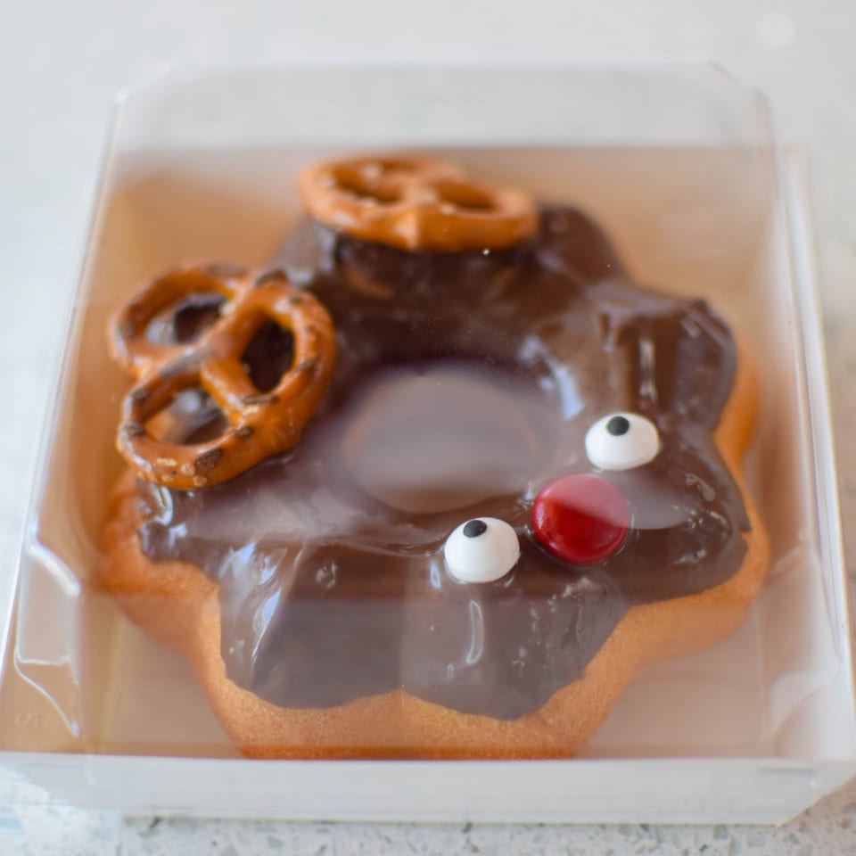 A monster-shaped donut with chocolate icing, pretzel ears, and candy eyes in a plastic container.