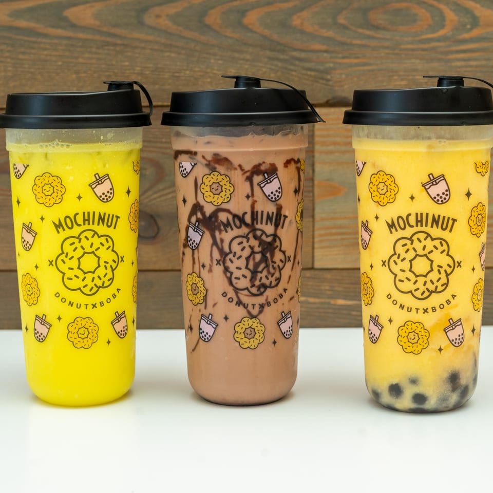 Three Mochinut drinks, two yellow and one chocolate, on a white surface with a wooden background.