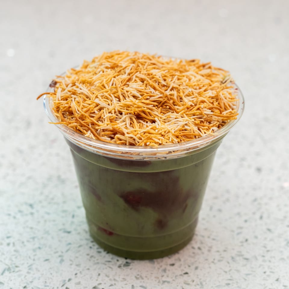 Green smoothie with red swirls, topped with crispy golden noodles in a clear plastic cup.