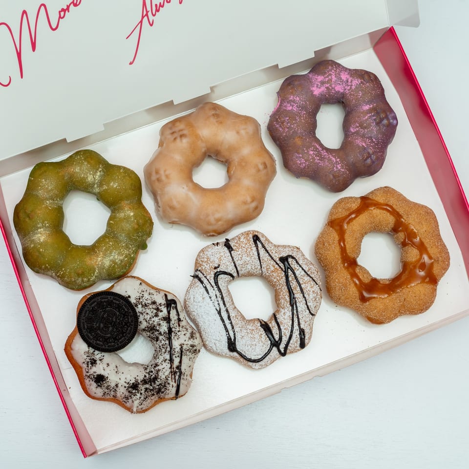 Six colorful, uniquely shaped donuts with various toppings in a white and pink box.