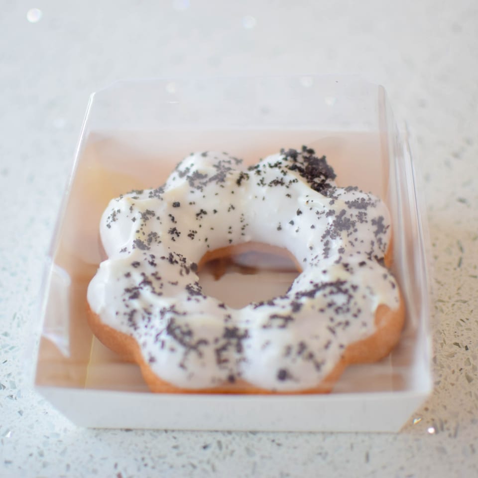 A flower-shaped mochi donut with white icing and dark sprinkles in a clear plastic container.