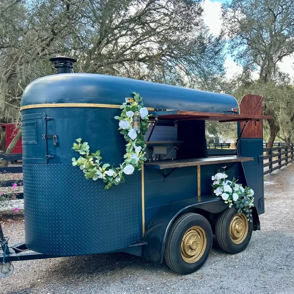 A dark blue food trailer with gold trim and wheels, decorated with white flowers, parked on a gravel path.