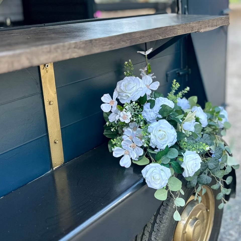 White roses and greenery adorn a dark blue food truck with a wooden counter and gold accents.