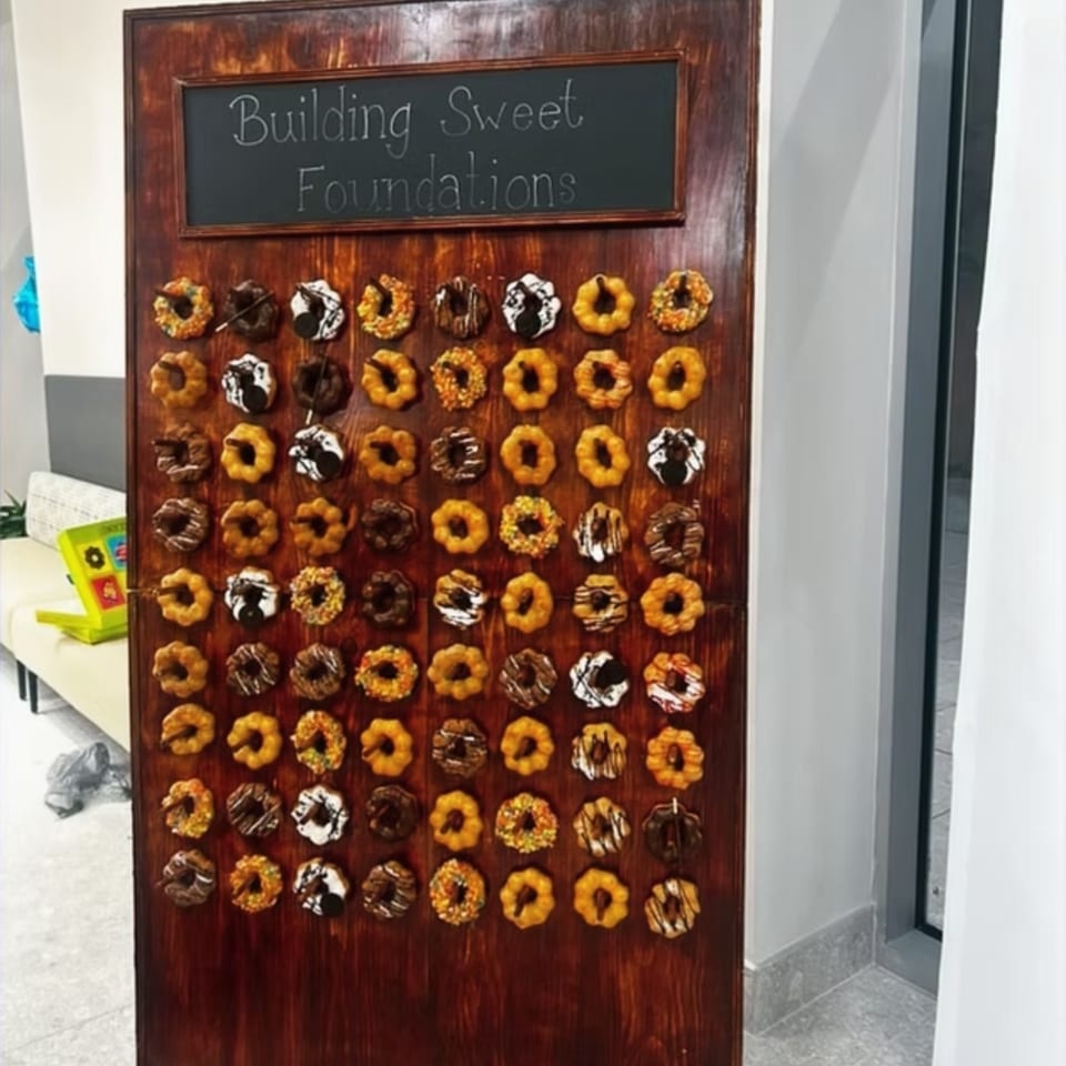 Wooden display board decorated with rows of assorted colorful donuts beneath a chalkboard reading “Building Sweet Foundations.”