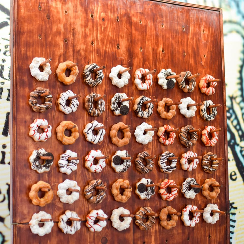 Rows of assorted glazed and powdered donuts, decorated with chocolate, red icing, and cookie pieces, arranged on a wooden board.