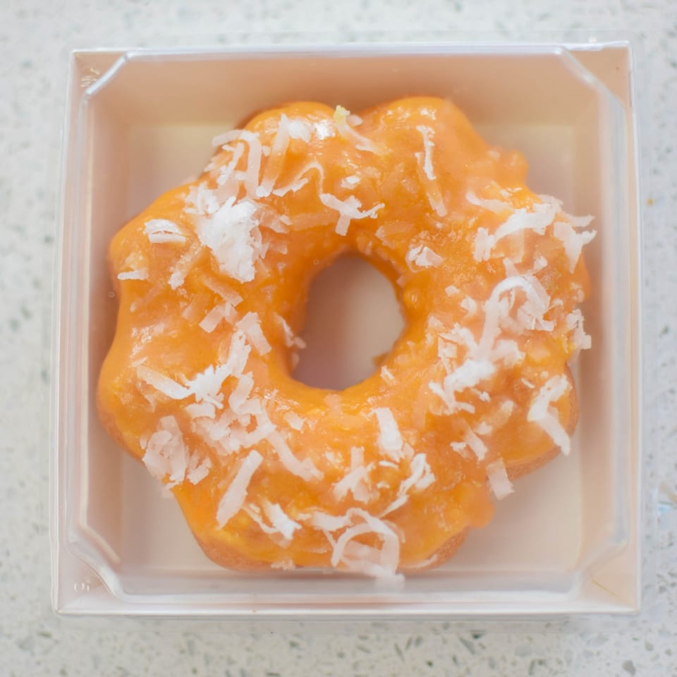 Orange donut with white shredded coconut flakes in a clear plastic container.