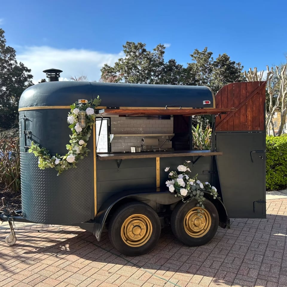 A dark blue food truck with gold accents and white floral decorations sits on a paved area.