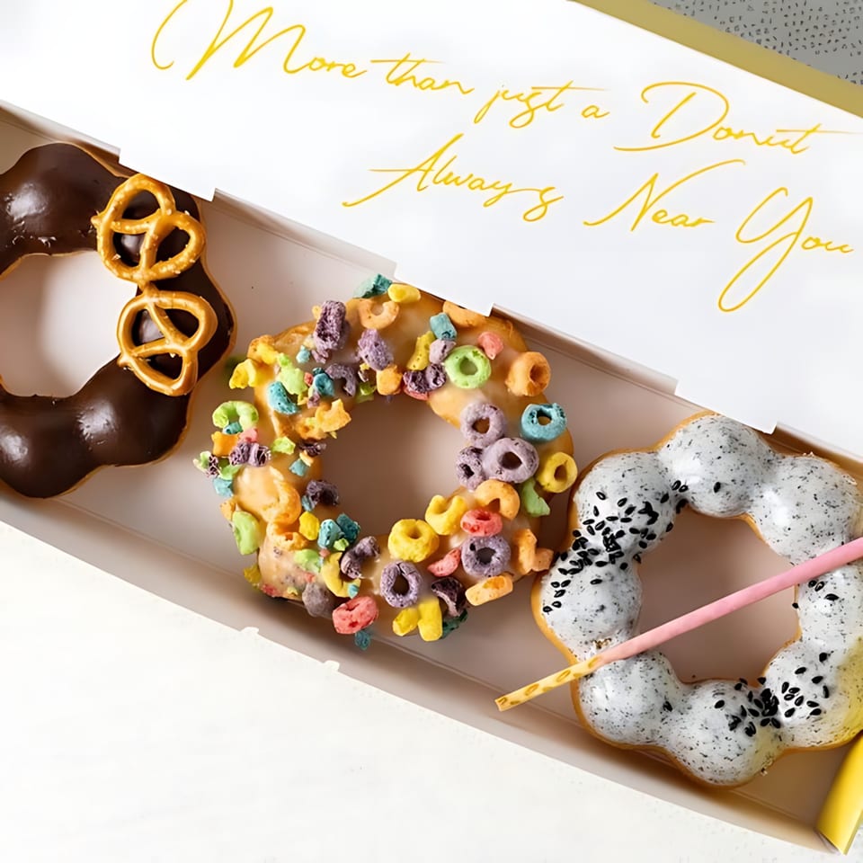 Three colorful mochi donuts, including chocolate pretzel, cereal, and black sesame, in a white box.