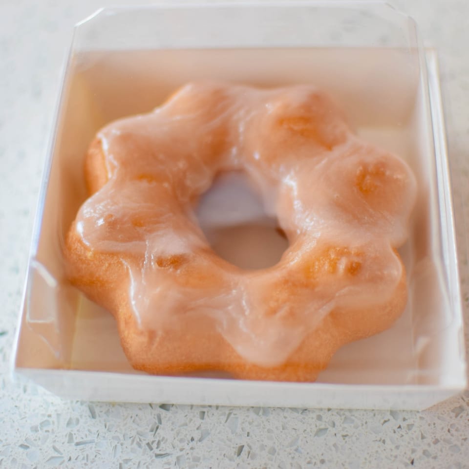 A glazed mochi donut in a clear plastic container, resting on a speckled white surface.