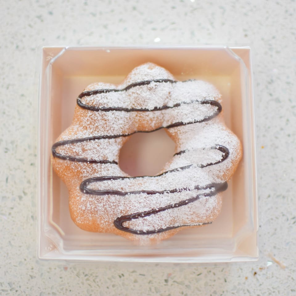 A star-shaped donut with powdered sugar and chocolate drizzle sits in a clear plastic container.