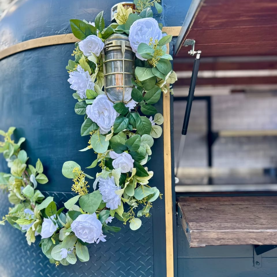 A dark blue food truck with a gold trim, decorated with a white floral garland and a rustic lantern.
