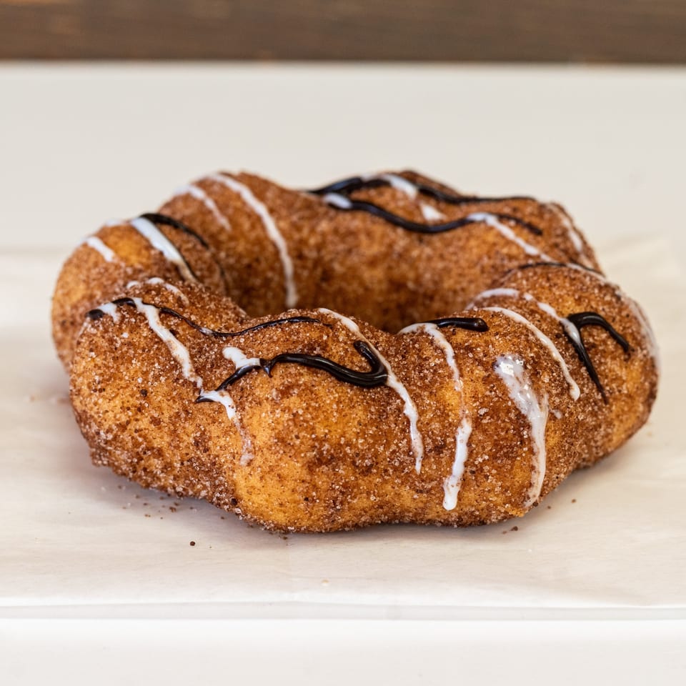 A churro donut, ring-shaped and covered in cinnamon sugar, with white and dark chocolate drizzles.