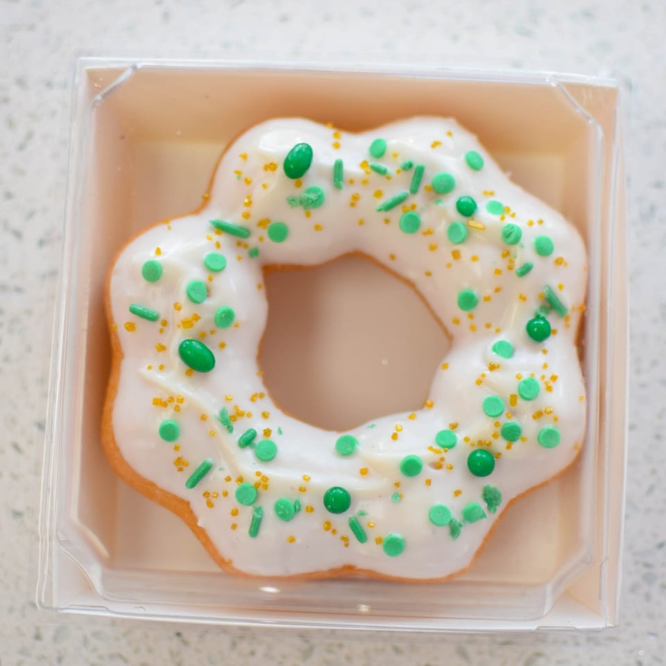 A mochi donut with white icing and green and gold sprinkles in a clear plastic container.