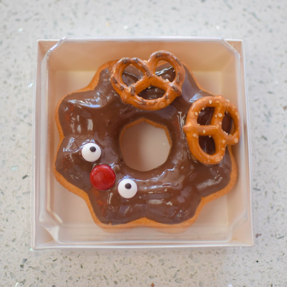 A reindeer-shaped donut with chocolate icing, pretzel antlers, candy eyes, and a red candy nose.