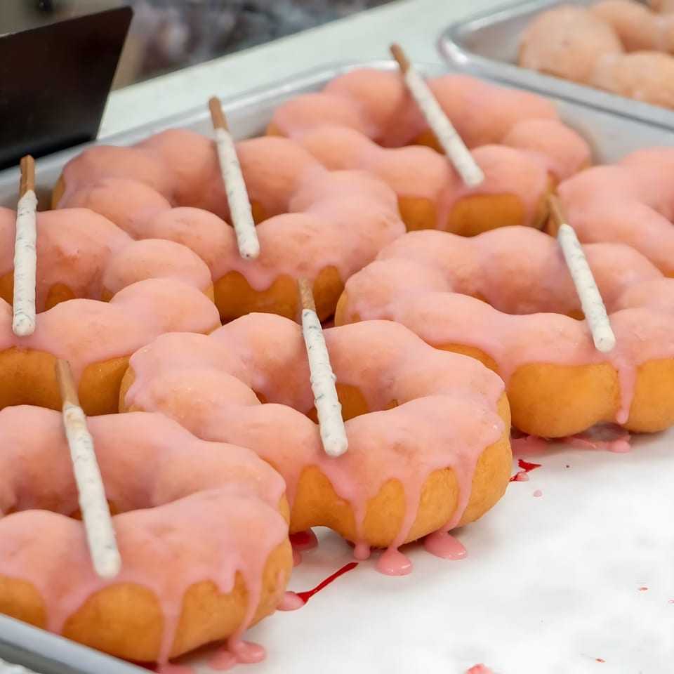 Rows of pink-glazed mochi donuts with white chocolate-dipped pretzel sticks on a tray.